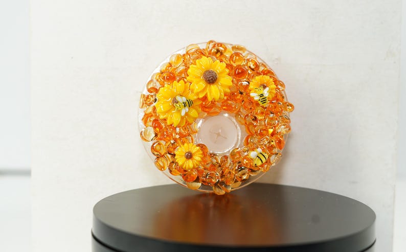 May include: A decorative candle holder with a clear glass base, adorned with yellow sunflowers, orange beads, and small bee figurines. The candle holder is designed to hold a single tea light.