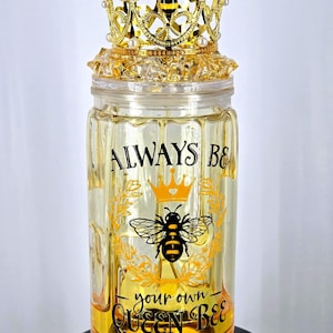 May include: A decorative glass jar with a gold-colored crown lid. The jar features the text "ALWAYS BE your own QUEEN BEE" with a bee and crown graphic. The crown is gold with faux pearls and a bee accent.