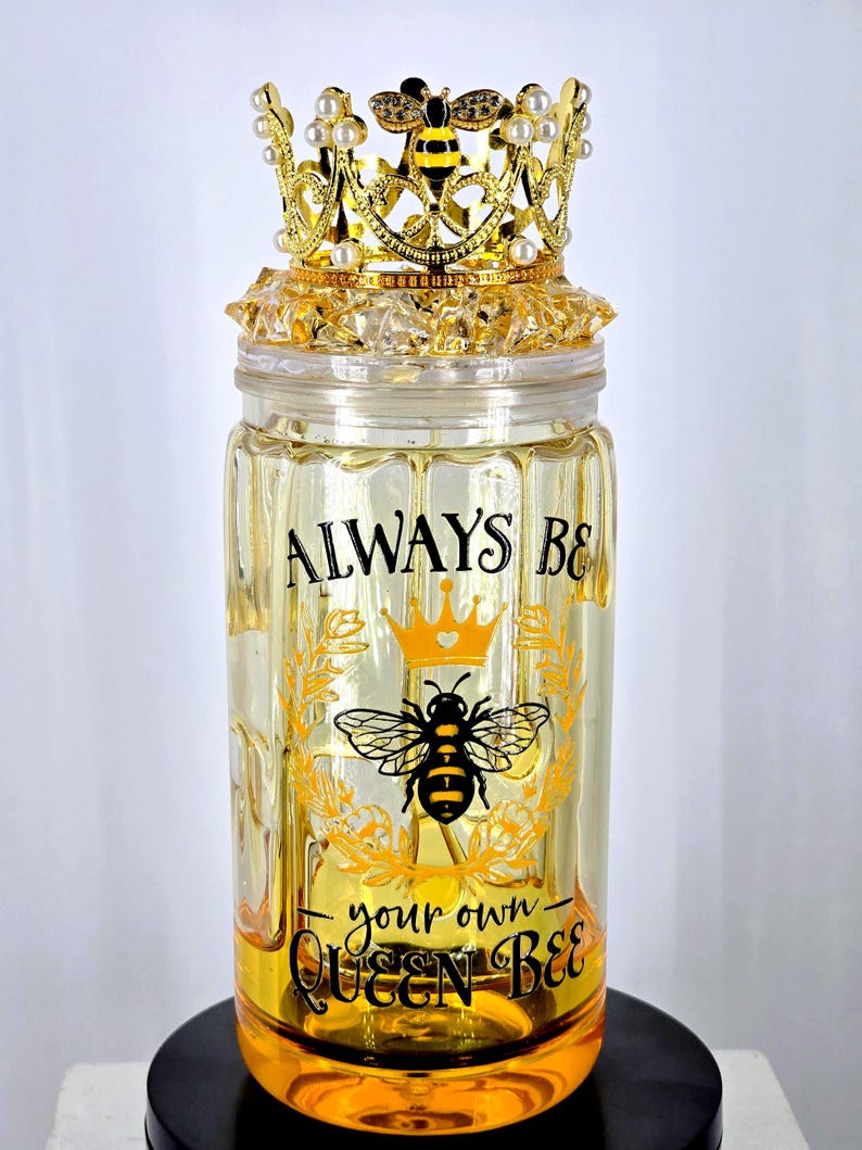 May include: A decorative glass jar with a gold-colored crown lid. The jar features the text "ALWAYS BE your own QUEEN BEE" with a bee and crown graphic. The crown is gold with faux pearls and a bee accent.
