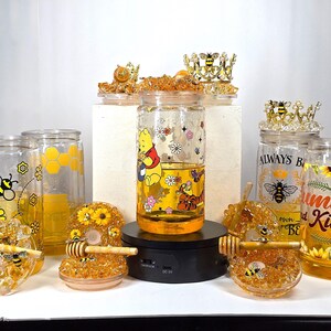 May include: A collection of glass jars filled with a golden liquid, possibly honey, featuring bee and Winnie the Pooh designs. Decorative honey dippers and bee-themed jar toppers are also present, creating a cohesive theme.