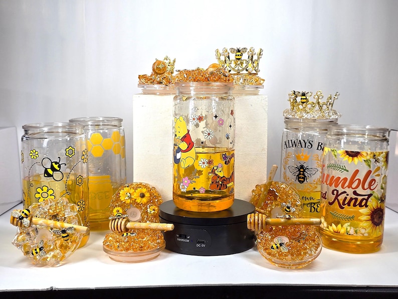 May include: A collection of glass jars filled with a golden liquid, possibly honey, featuring bee and Winnie the Pooh designs. Decorative honey dippers and bee-themed jar toppers are also present, creating a cohesive theme.