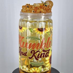 May include: A clear glass jar with a decorative lid featuring a honey and bee design. The jar is adorned with sunflowers and the words "Humble And Kind" in orange and burgundy lettering. The jar is sitting on a black stand.
