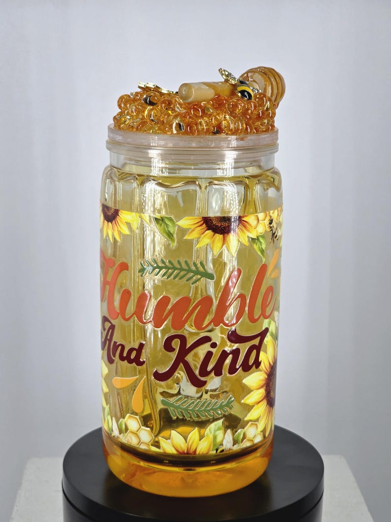 May include: A clear glass jar with a decorative lid featuring a honey and bee design. The jar is adorned with sunflowers and the words "Humble And Kind" in orange and burgundy lettering. The jar is sitting on a black stand.