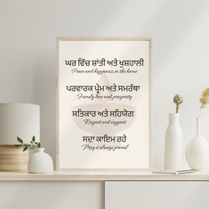 Może przedstawiać: Wydruk w ramie z białym tłem i czarnym tekstem w języku pendżabskim. Tekst tłumaczy się jako "Spokój i szczęście w domu, Miłość rodzinna i dobrobyt, Szacunek i wsparcie, Niech zawsze trwa". Wydruk przedstawia stylizowane zdjęcie zapalanej diya, tradycyjnej indyjskiej lampy naftowej.