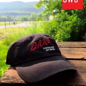 Peut inclure: Casquette de baseball noire avec un texte brodé en rouge qui dit "DARE KEEPING KIDS OFF DRUGS."