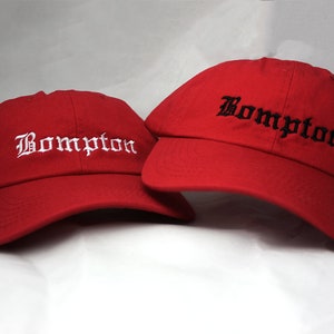 May include: Two red baseball caps with black stitching. One cap has the word "Compton" embroidered in white, and the other has the word "Compton" embroidered in black.