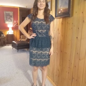Puede incluir: Una mujer con un vestido de encaje azul verdoso con un dobladillo festoneado. Est&aacute; de pie frente a una pared de madera y lleva sandalias negras.