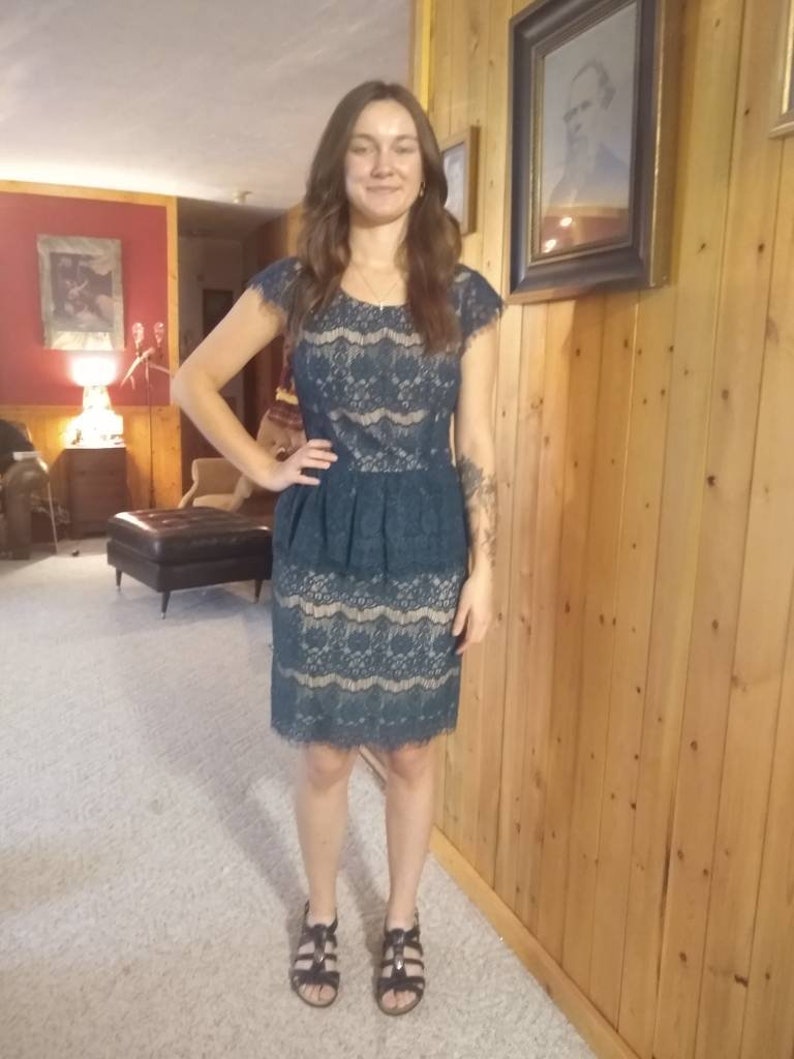 Puede incluir: Una mujer con un vestido de encaje azul verdoso con un dobladillo festoneado. Est&aacute; de pie frente a una pared de madera y lleva sandalias negras.