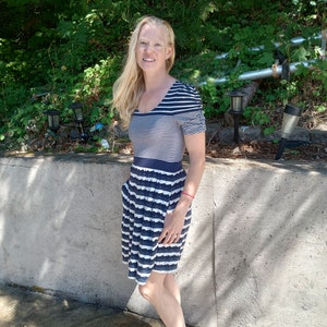 May include: A navy blue and white striped dress with short sleeves and a pleated skirt. The dress has a scoop neckline and pockets. The woman is standing outside in front of a concrete wall and greenery.