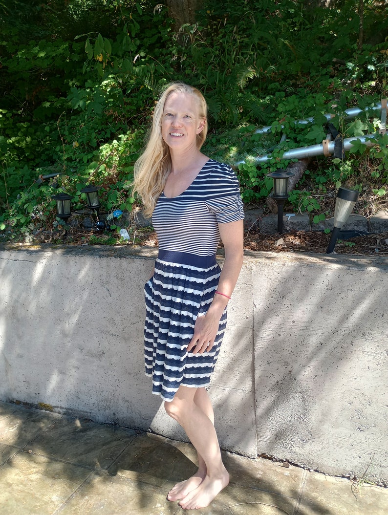 May include: A navy blue and white striped dress with short sleeves and a pleated skirt. The dress has a scoop neckline and pockets. The woman is standing outside in front of a concrete wall and greenery.