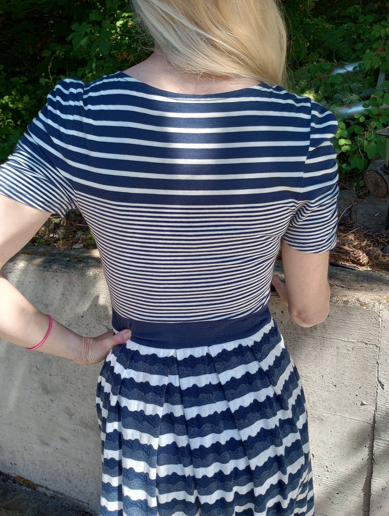 May include: A navy and white striped dress. The top has short sleeves and a scoop neckline. The skirt is pleated with a scalloped edge. The dress is fitted at the waist.