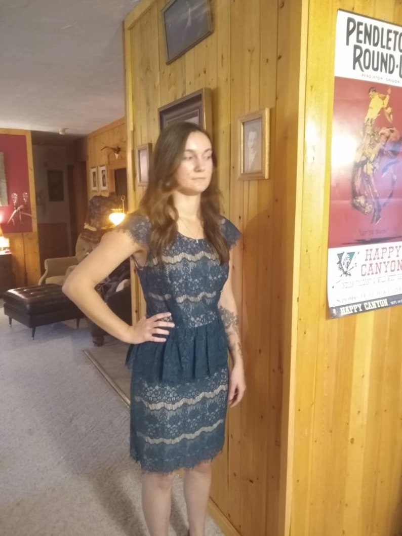 Puede incluir: Una mujer lleva un vestido de encaje azul turquesa con una cintura peplum con volantes. El vestido tiene un corpi&ntilde;o ajustado y una falda que le llega a las rodillas.