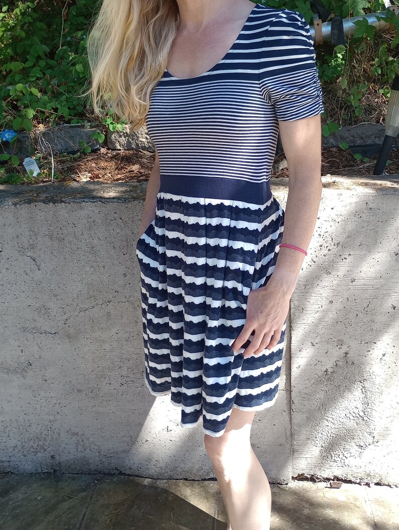 May include: A navy and white striped dress with a scoop neckline and short sleeves. The dress has a fitted bodice and a flared skirt with pockets. The skirt has a scalloped hemline. The dress is made of a soft, lightweight fabric.