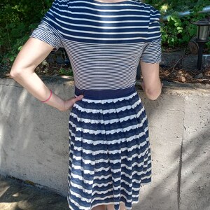 May include: A navy and white striped dress with a fitted bodice and a pleated skirt. The dress has short sleeves and a navy waistband. The skirt has a scalloped edge. The dress is knee-length.