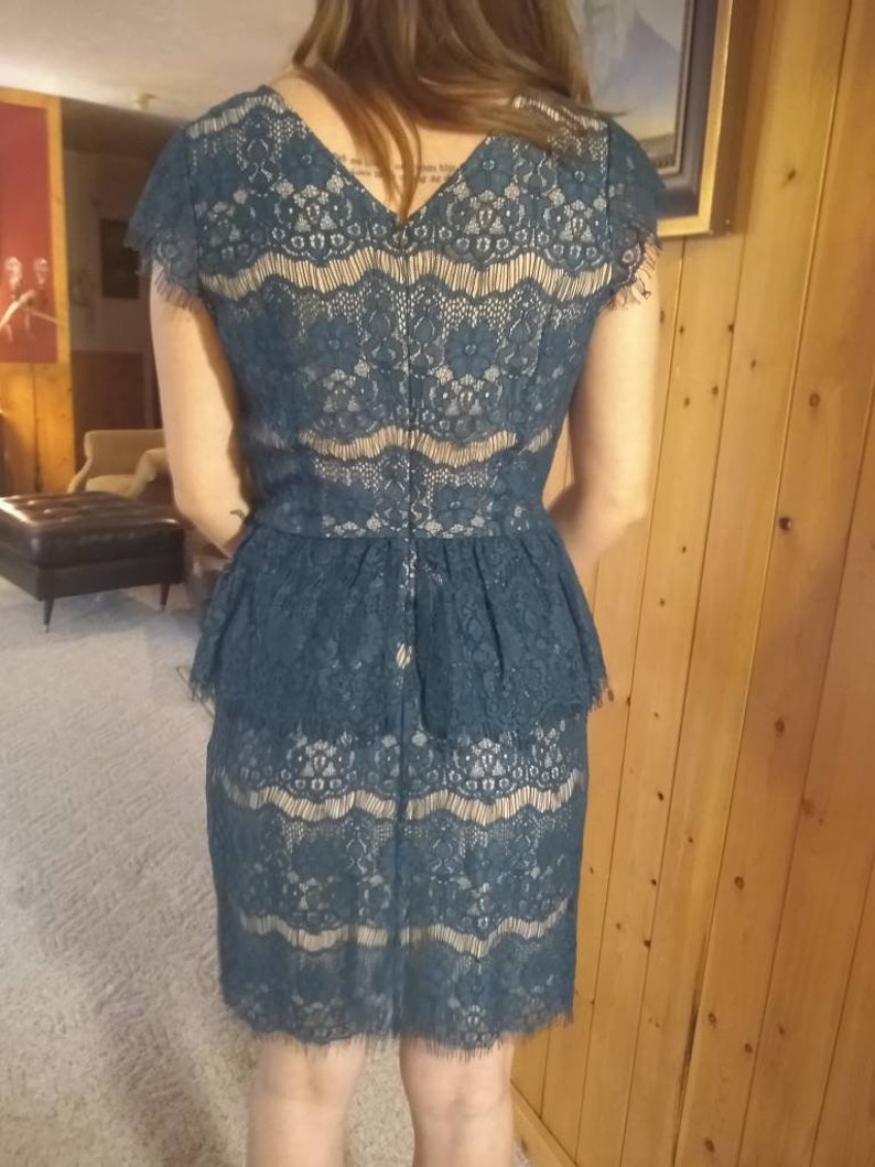 Puede incluir: Un vestido de encaje azul teal con un corpi&ntilde;o ajustado y una falda acampanada. El vestido tiene un escote en V y mangas cortas.