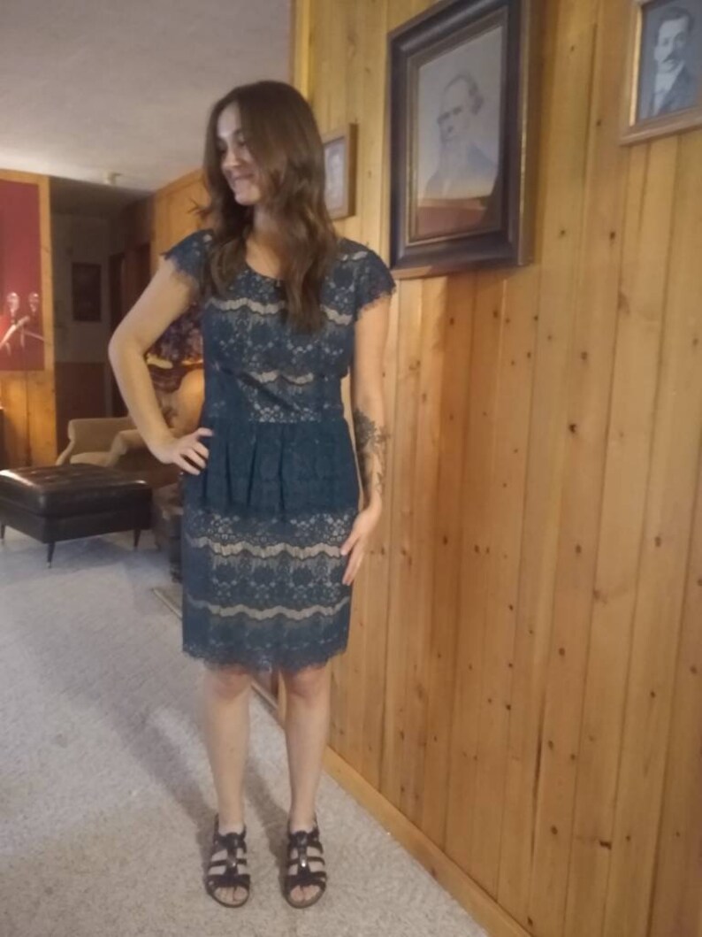Puede incluir: Una mujer con un vestido de encaje azul verdoso con una falda con volantes. Est&aacute; de pie frente a una pared de madera.