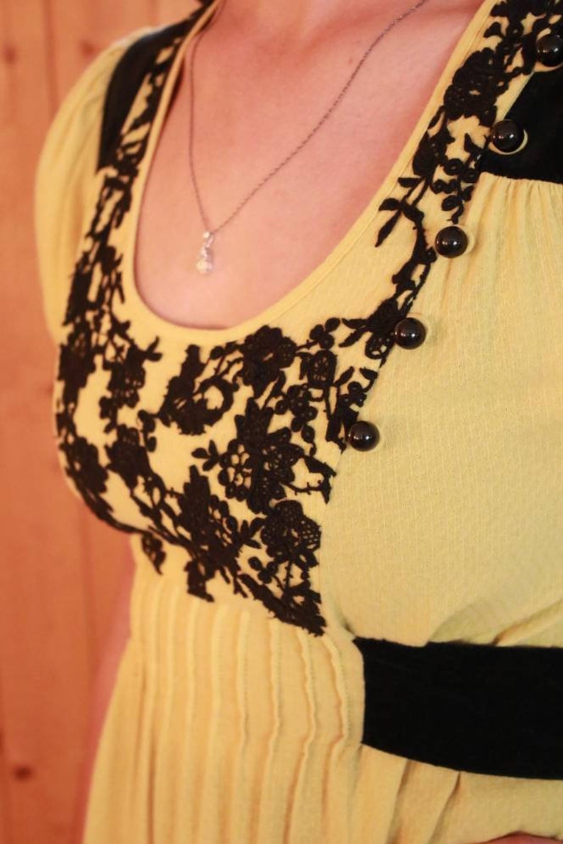Op de afbeelding: Een gele jurk met zwarte kanten details en zwarte knopen. De jurk heeft een ronde halslijn en een zwarte band in de taille. Een zilveren ketting met een kleine hanger is zichtbaar.