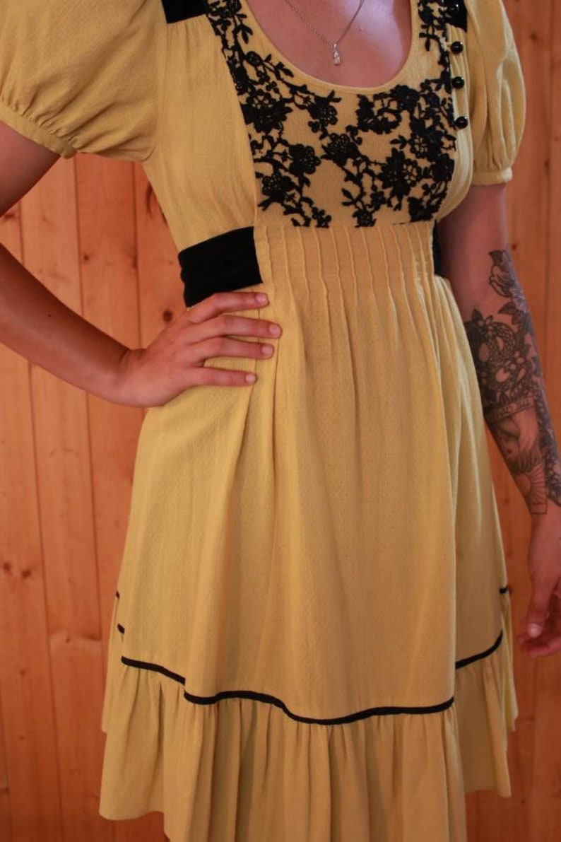 Op de afbeelding: Mosterdgele jurk met zwarte kanten details en zwarte bies. De jurk heeft korte, gepofte mouwen, een getailleerde taille en een geplooide rok. De jurk heeft een vintage uitstraling.
