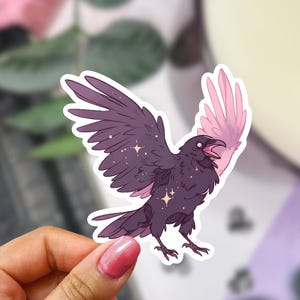 Op de afbeelding: Een sticker met een gestileerde raaf met uitgespreide vleugels. De raaf is voornamelijk donkerpaars met roze accenten op één vleugel. Het ontwerp bevat sprankelende sterdetails, wat een grillige esthetiek creëert.