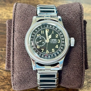 May include: An Oris automatic wristwatch with a black dial, silver-tone hands, and Arabic numerals. The watch features a date pointer hand, a small seconds sub-dial, and a coin-edge bezel. It is attached to a silver-tone metal link bracelet.