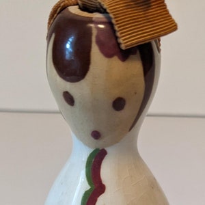 May include: A ceramic doll figurine with a brown and white painted face, brown hair, and a brown bow. The doll is wearing a white dress with green and red floral designs. The doll is standing on a white surface.