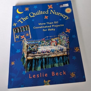May include: The Quilted Nursery book cover features a blue background with stars and a cot with a quilted bedding set. The title of the book is "The Quilted Nursery" and the subtitle is "More Than 50 Coordinated Projects for Baby". The author's name is Leslie Beck.