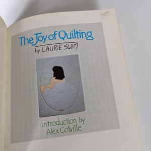 Pode incluir: The Joy of Quilting de Laurie Swim, uma capa de livro com um design acolchoado branco de uma mulher com um vestido branco e um laço rosa.