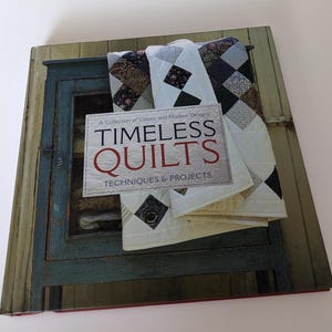 Puede incluir: Un libro titulado "Timeless Quilts: Techniques & Projects" con una colección de diseños clásicos y modernos. La portada muestra una pila de colchas con varios patrones en negro, blanco y gris.