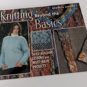 May include: A book titled "Knitting Beyond the Basics" by Rebecca Lennox. The cover features a woman wearing a blue knitted sweater, a close-up of a blue and black knitted fabric, and a brown knitted sweater with a colorful detail.
