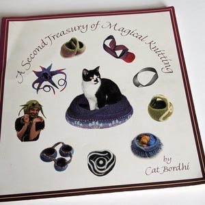 May include: A book cover with the title "A Second Treasury of Magical Knitting" by Cat Bordhi. The cover features a black and white cat sitting in a knitted cat bed, surrounded by various knitted items, including a hat, a pair of slippers, a basket, and a bowl.