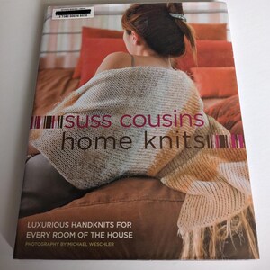 May include: A book titled "Suss Cousins Home Knits" with a photo of a woman wearing a knitted throw on a couch. The book cover text reads "Luxurious Handknits for Every Room of the House" and "Photography by Michael Weschler".