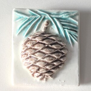 May include: A rectangular ceramic tile featuring a detailed relief of a pine cone. The pine cone is brown with textured scales, set against a white background. Above the pine cone are stylized turquoise pine needles. The tile is likely for decorative use.