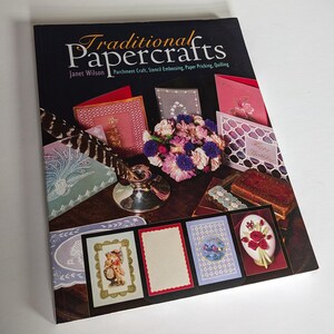 May include: A book titled "Traditional Papercrafts" by Janet Wilson. The book cover features various paper crafts, including a card with the text "Happy Birthday", a card with a floral design, and a card with a feather design. The book cover also features a bouquet of pink and purple flowers.