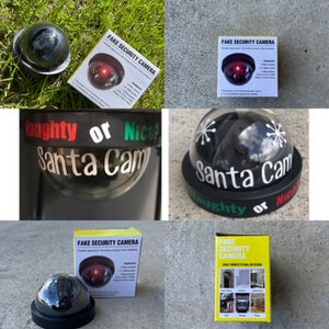 May include: A fake security camera with a red LED light and a dome-shaped lens. The camera is labeled "Fake Security Camera" and has a sticker that says "Naughty or Nice? Santa Cam".