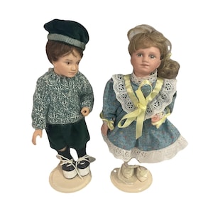 May include: Two porcelain dolls, a boy and a girl, stand on cream-colored bases. The boy doll wears a green sweater, shorts, and a cap. The girl doll is in a blue floral dress with lace trim and a yellow ribbon.