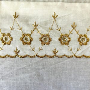 May include: Close-up of white fabric with a detailed gold embroidered floral border. The embroidery features repeating flower and leaf patterns, creating a decorative trim. The fabric has a subtle texture.