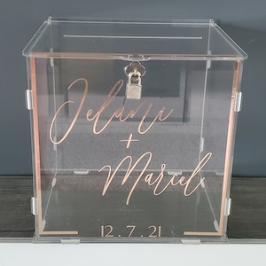 May include: Clear acrylic wedding card box with a rose gold trim and lock. The box has the names "Jelena + Marcel" and the date "12.7.21" written on it in rose gold lettering.