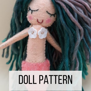 Può includere: Una bambola di sirena in feltro con lunghi capelli in lana turchese e viola, bottoni bianchi per la parte superiore e una coda rosa. La bambola sorride con gli occhi chiusi. Il testo "DOLL PATTERN" è stampato su uno striscione bianco sotto la bambola.