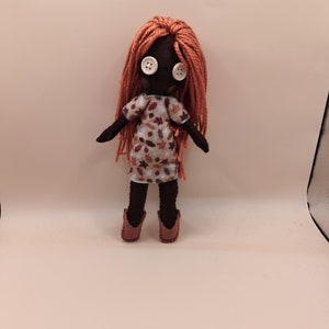 May include: A handmade doll with a dark complexion, button eyes, and long orange yarn hair. The doll wears a patterned dress with brown and beige leaf designs and matching brown boots. The doll's arms are outstretched.