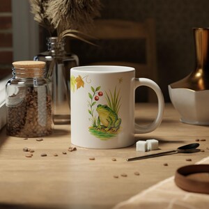 May include: White ceramic mug featuring a green frog illustration with red berries and tall grass. The mug has a curved handle and is decorated with autumn leaves. The mug is on a wooden surface with coffee beans and a spoon.