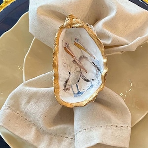 May include: A decorative oyster shell napkin ring with a gold-painted rim, featuring a watercolor painting of two pelicans. The napkin ring is set on a folded beige linen napkin, atop a cream-colored plate, with a navy blue charger.