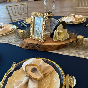 Può includere: Elegante mise en place con tovaglia blu navy, piatti bordati d'oro e posate dorate. Una cornice dorata, una lampada a olio in vetro trasparente e un grande fiore bianco sono su un centrotavola in legno. I tovaglioli sono adornati da anelli portatovaglioli a conchiglia.