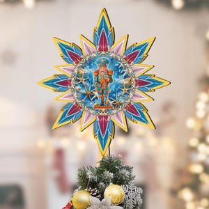 May include: A star-shaped Christmas tree topper with a detailed illustration of a nutcracker soldier. The topper features a circular center with the nutcracker and a starburst design in blue, yellow, pink, and red. The topper is placed on top of a decorated Christmas tree.