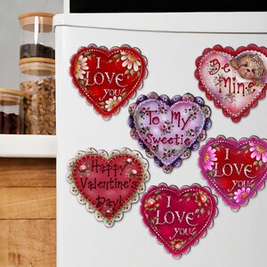 May include: Six heart-shaped magnets with various Valentine's Day messages. The magnets are decorated with flowers, glitter, and other embellishments. The messages include "I Love You", "To My Sweetie", "Happy Valentine's Day", "Be Mine", and "I Love You".