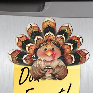 May include: A painted wooden turkey magnet with a brown, black, and orange patterned tail. The turkey is holding a small chick wearing a black hat. The magnet is attached to a yellow sticky note with the text "Don't Forget!"