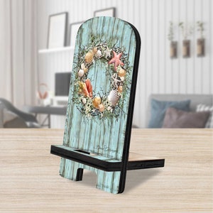 May include: A teal blue wooden phone stand with a seashell wreath design. The stand has a black metal kickstand and a white inscription that reads "Dana DeSena".