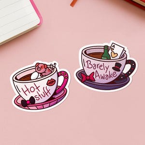 Könnte beinhalten: Zwei skurrile Aufkleber mit Kaffeetassen. Einer sagt "Hot stuff" und hat ein Schwein und ein Ei darin. Der andere sagt "Barely Awake" und hat eine Flasche und eine Spielkarte. Die Aufkleber befinden sich auf einer rosa Oberfläche mit einem Notizbuch und einem Journal.