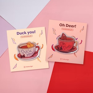 Könnte beinhalten: Zwei Emaille-Anstecknadeln auf Karten. Eine Nadel sagt "Duck you!" mit einer Ente in einer Teetasse. Die andere sagt "Oh Deer!" mit einem Teetassen-Design. Beide Nadeln befinden sich auf pfirsichfarbenen Karten mit geometrischen Formen.