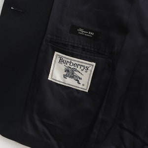May include: A close-up of the inside of a black blazer. The dark lining reveals a pocket with a Burberrys label. A smaller label reads "Super 100 Finest Quality". The blazer is likely made of fine wool.