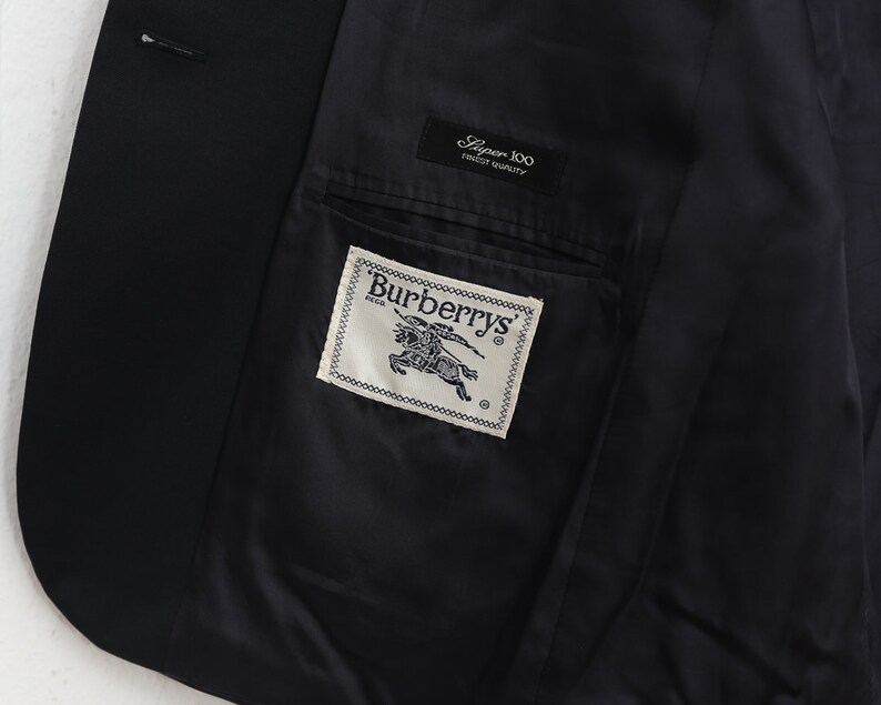 May include: A close-up of the inside of a black blazer. The dark lining reveals a pocket with a Burberrys label. A smaller label reads "Super 100 Finest Quality". The blazer is likely made of fine wool.